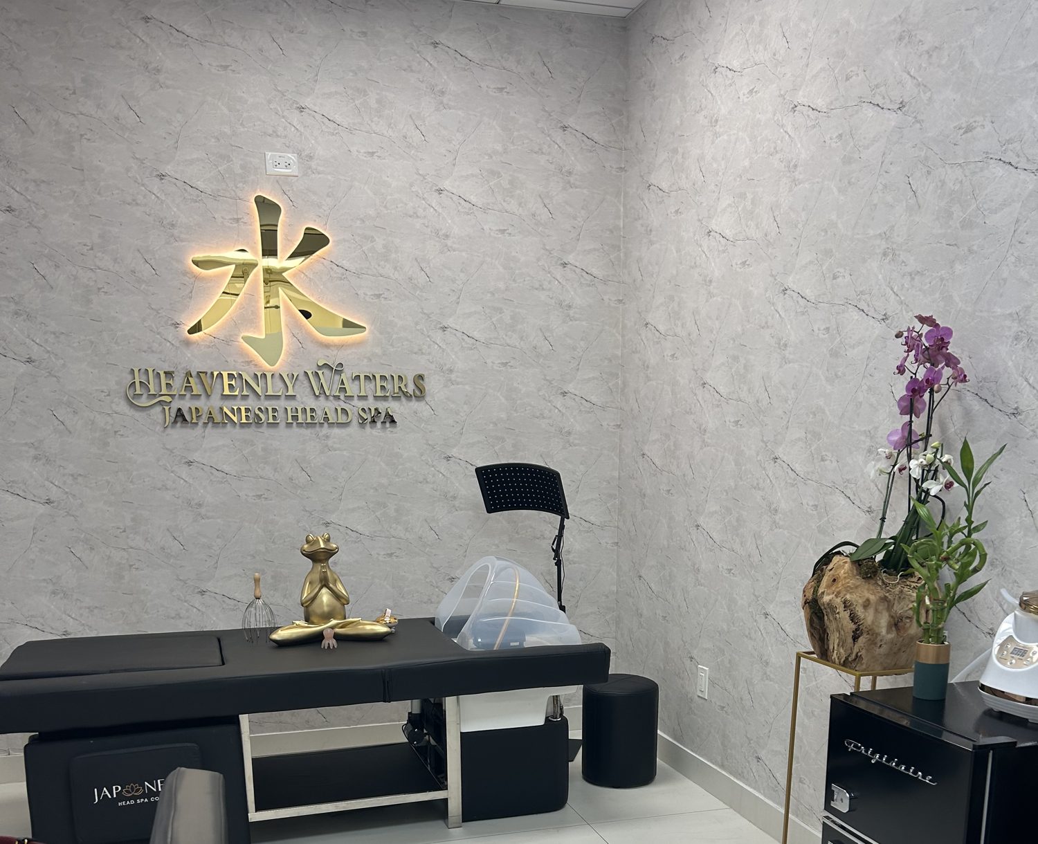 The actual Heavenly Waters studio with gold signage, marble-look walls, orchids, and a black heated head spa bed.